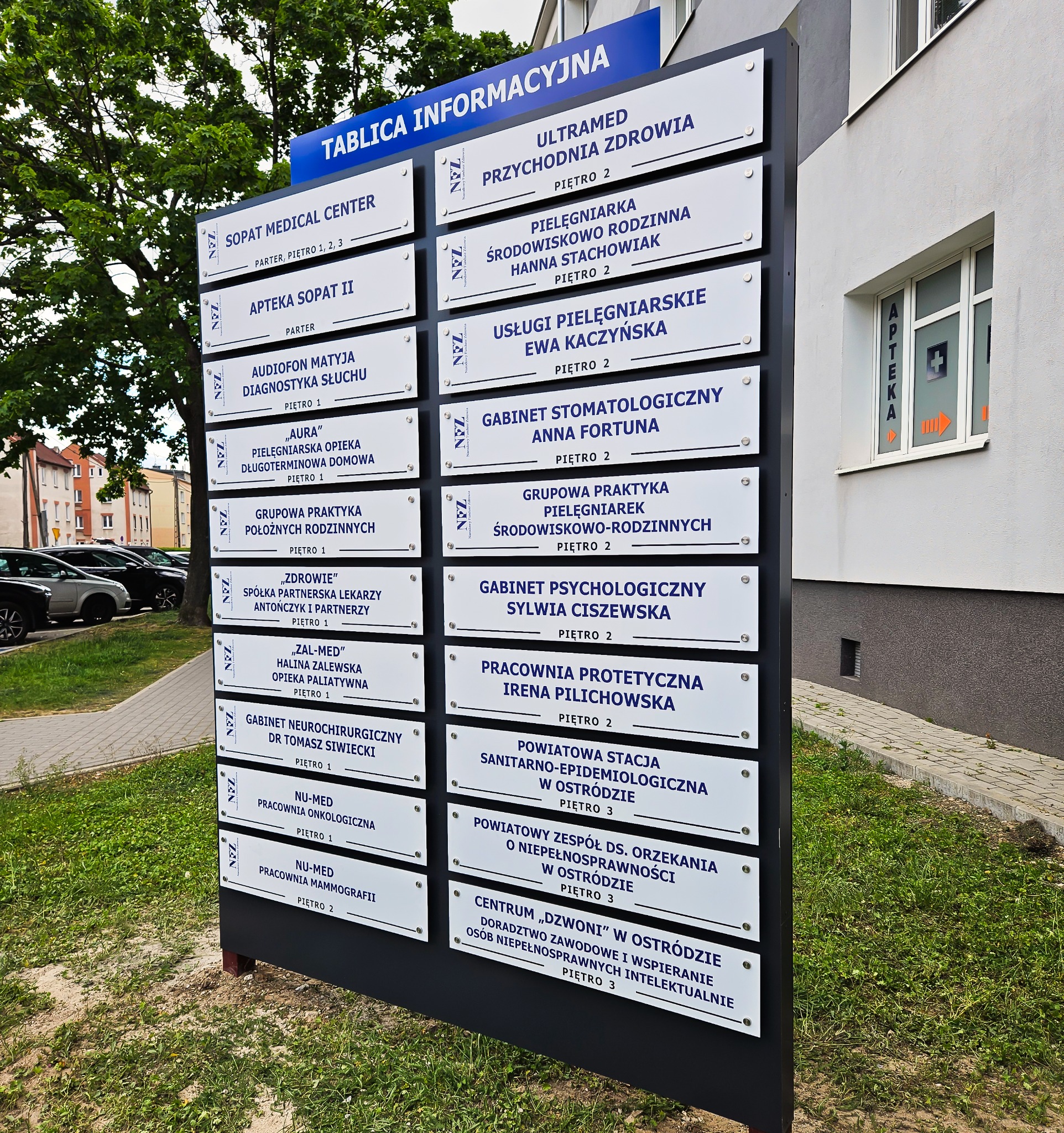 Tablica informacyjna z nazwami placówek medycznych i usługowych, umieszczona przed budynkiem. Widoczne nazwy firm i numery pięter.