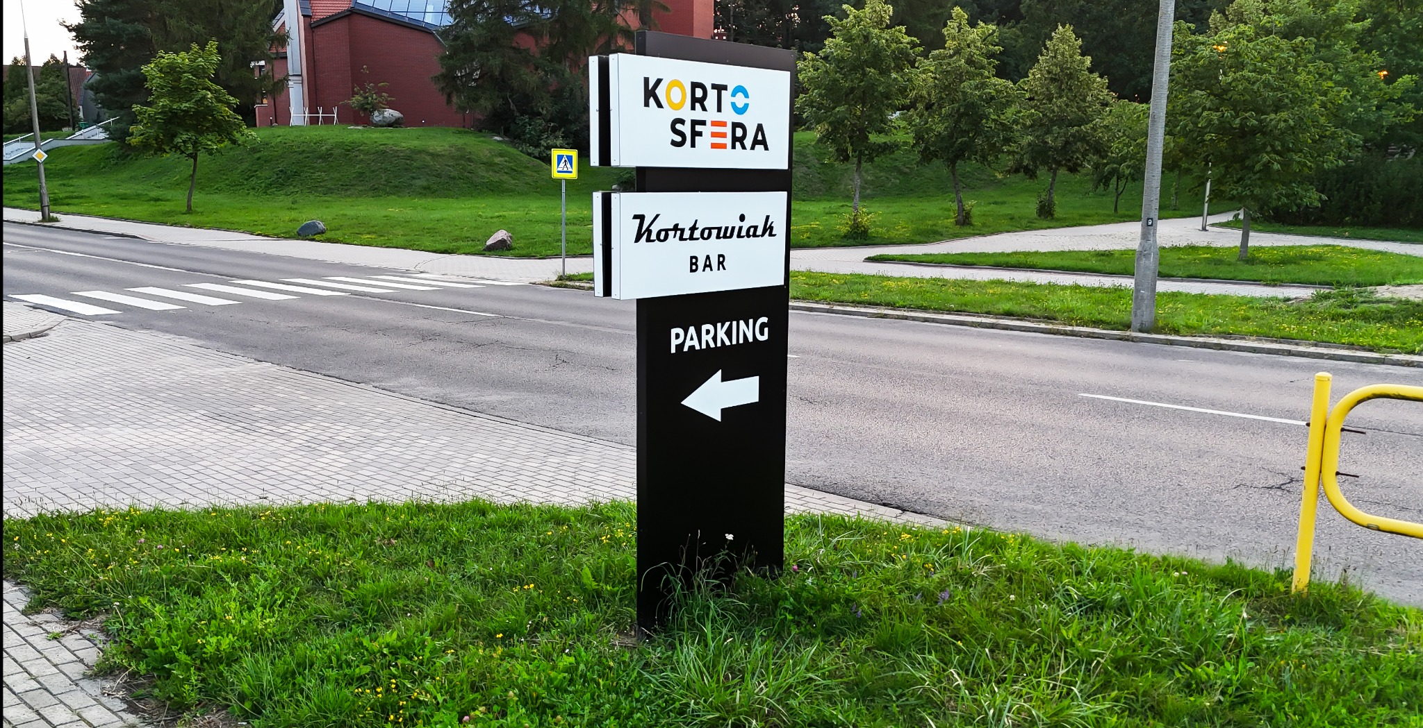 Czarny, wolnostojący pylon reklamowy z białymi napisami 'Kortosfera', 'Kortowiak Bar' i strzałką 'Parking', umieszczony przy ulicy i chodniku.