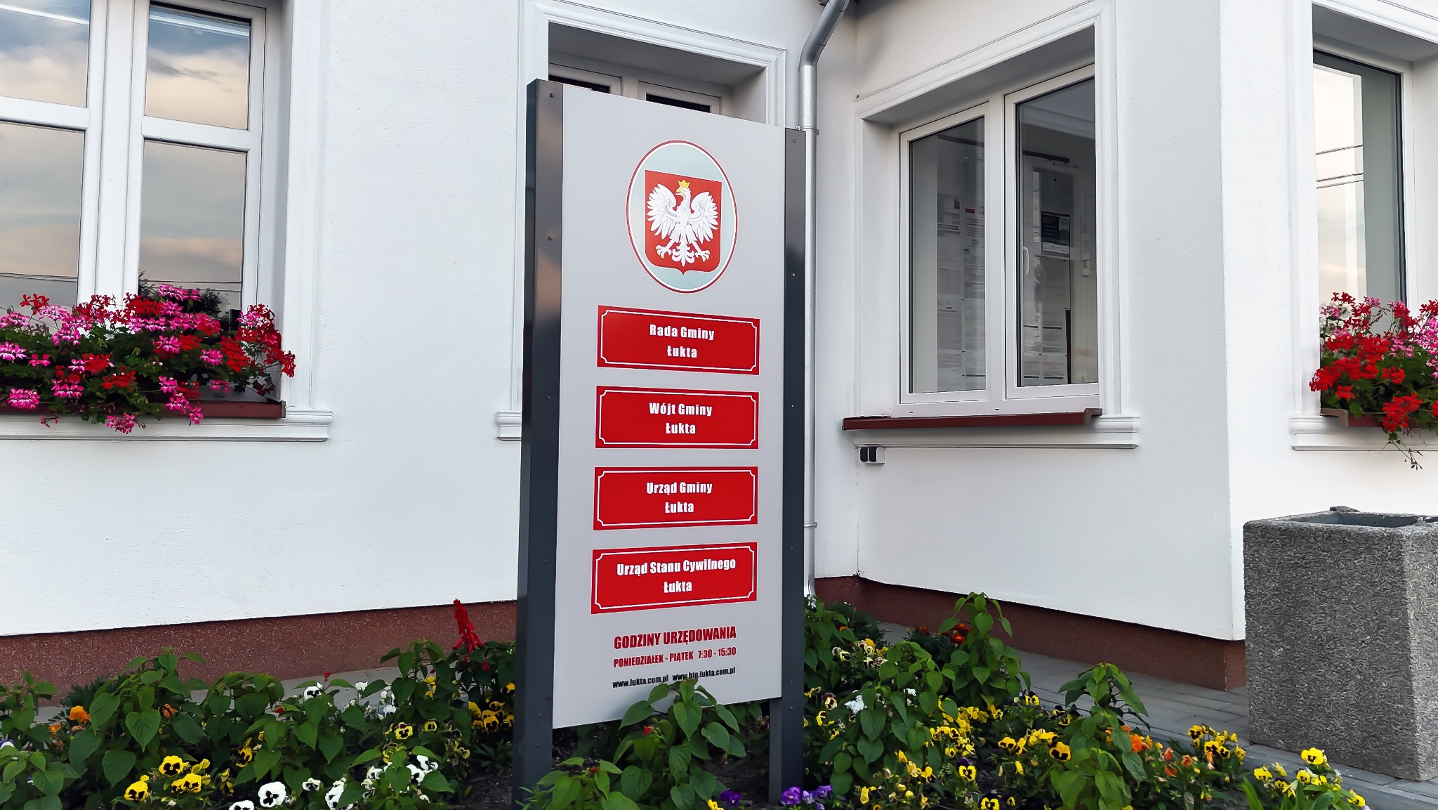 Tablica informacyjna przed budynkiem Urzędu Gminy Łukta z herbem Polski i nazwami wydziałów. W tle biała fasada budynku z oknami i kwiatami.