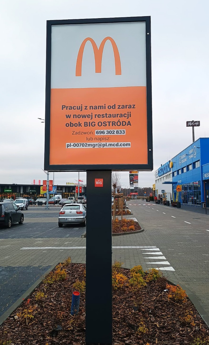 Billboard McDonald's przy parkingu centrum handlowego BIG Ostróda, zachęcający do pracy w nowej restauracji. Widoczne logo i dane kontaktowe w kolorze pomarańczowym.