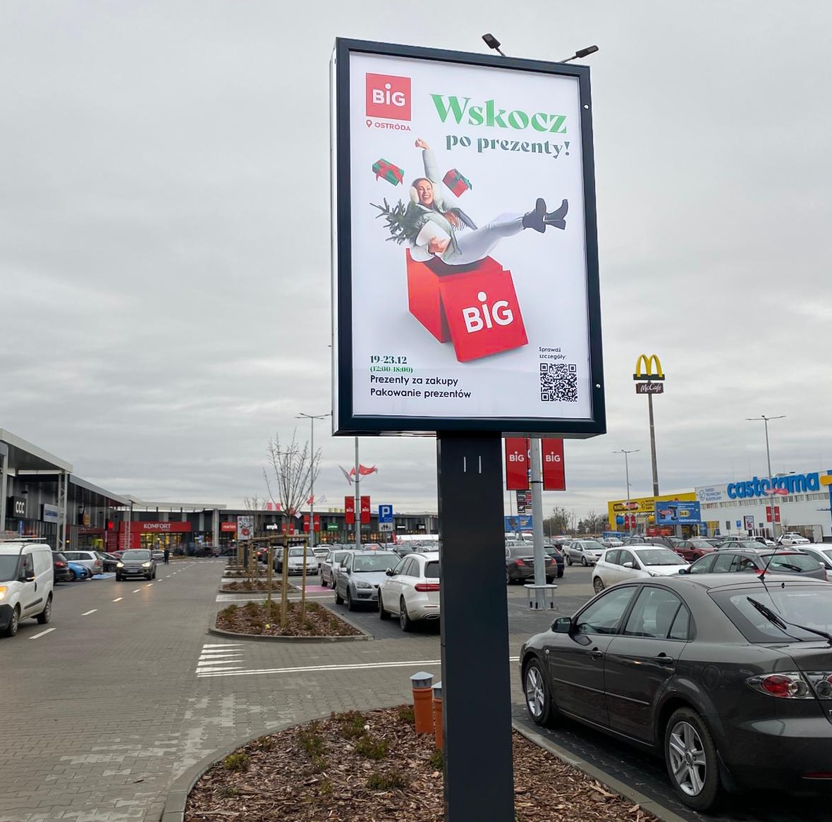 Billboard reklamowy sklepu BIG w Ostródzie. Na plakacie kobieta wpadająca do czerwonego pudełka z napisem BIG, hasło: Wskocz po prezenty!, data i kod QR.