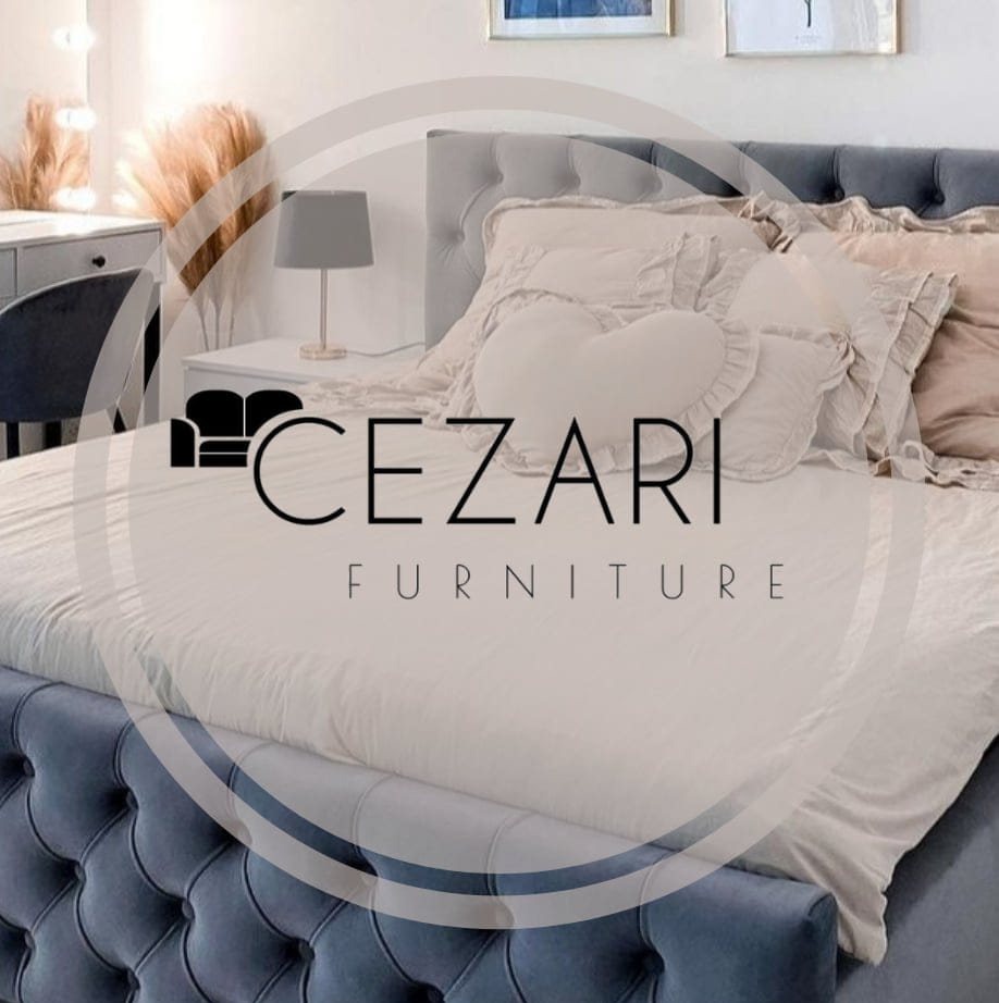 Tapicerowane łóżko z pikowanym zagłówkiem w szaro-niebieskim odcieniu, z dekoracyjnymi poduszkami i pościelą. W tle toaletka z oświetleniem i dekoracjami.