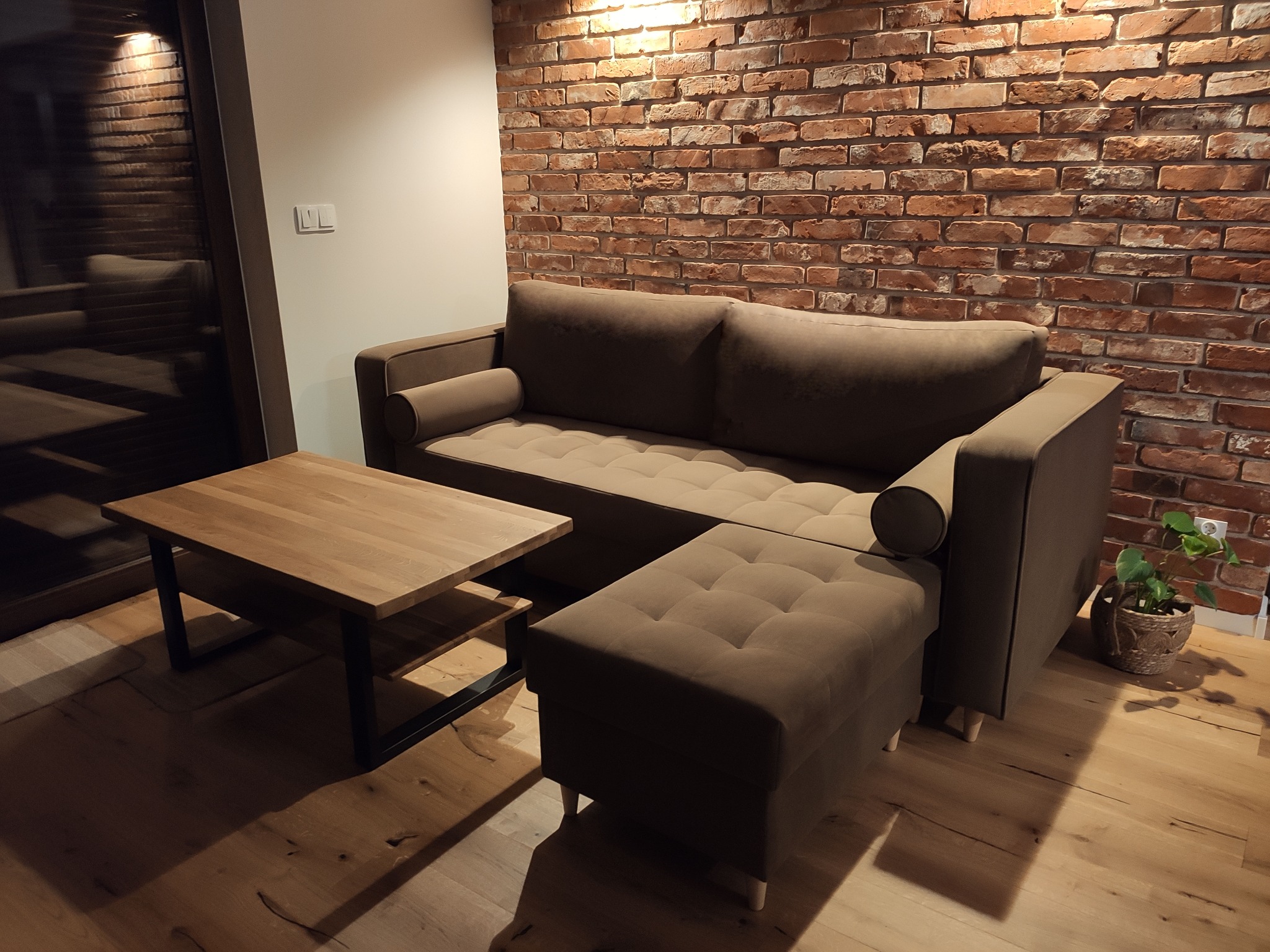 Beżowa sofa z pufą i drewnianym stolikiem kawowym na tle ściany z czerwonej cegły i drewnianej podłogi. W rogu widoczna roślina doniczkowa.