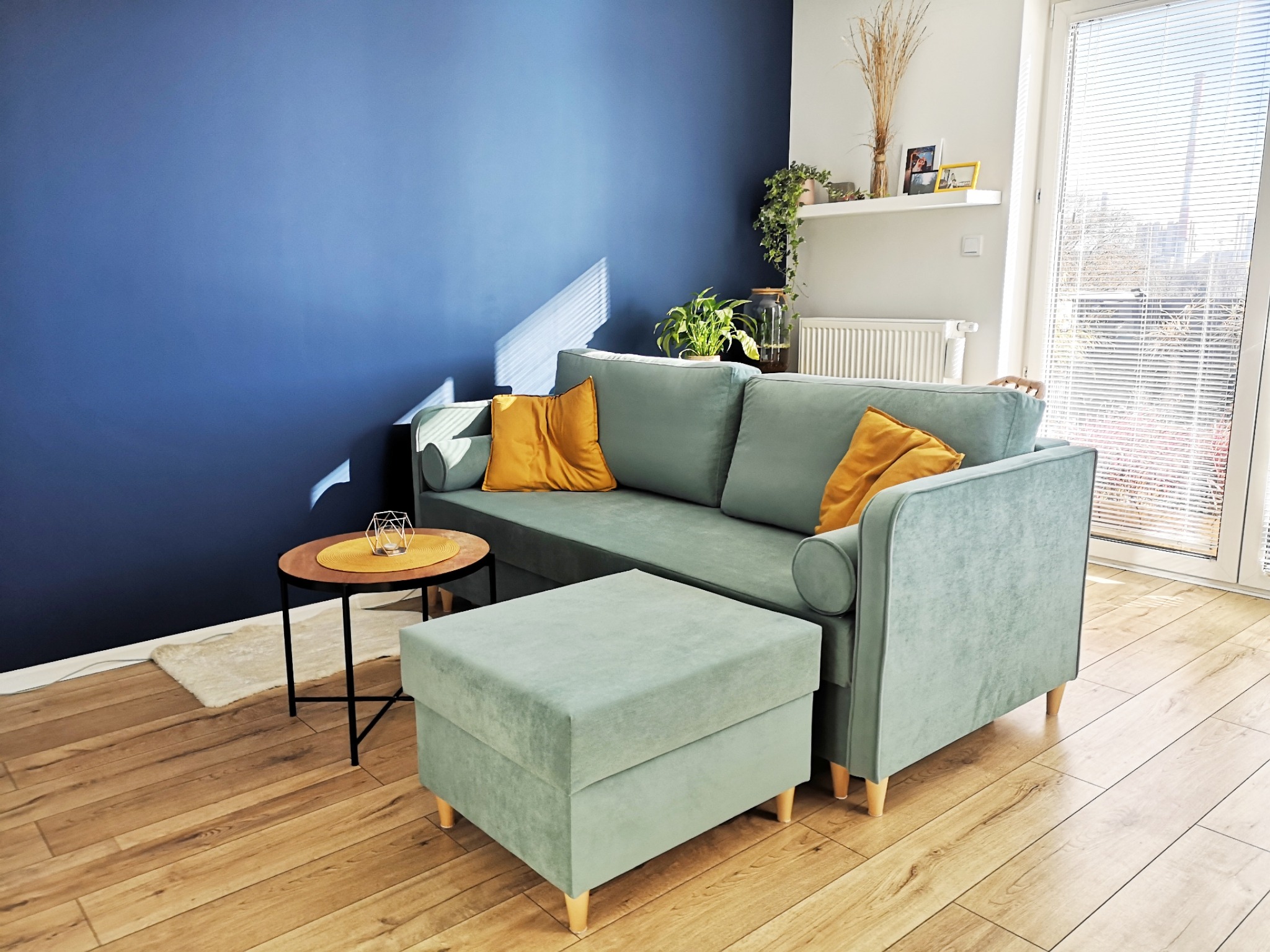 Turkusowa sofa z podnóżkiem i żółtymi poduszkami na tle granatowej ściany. Na stoliku dekoracja. Jasne drewniane panele, okno z żaluzjami.