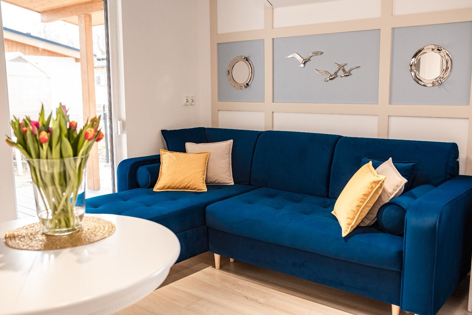 Niebieski narożnik z weluru z żółtymi poduszkami w salonie. Dekoracje ścienne w stylu marynistycznym. Na pierwszym planie biały stolik z tulipanami.
