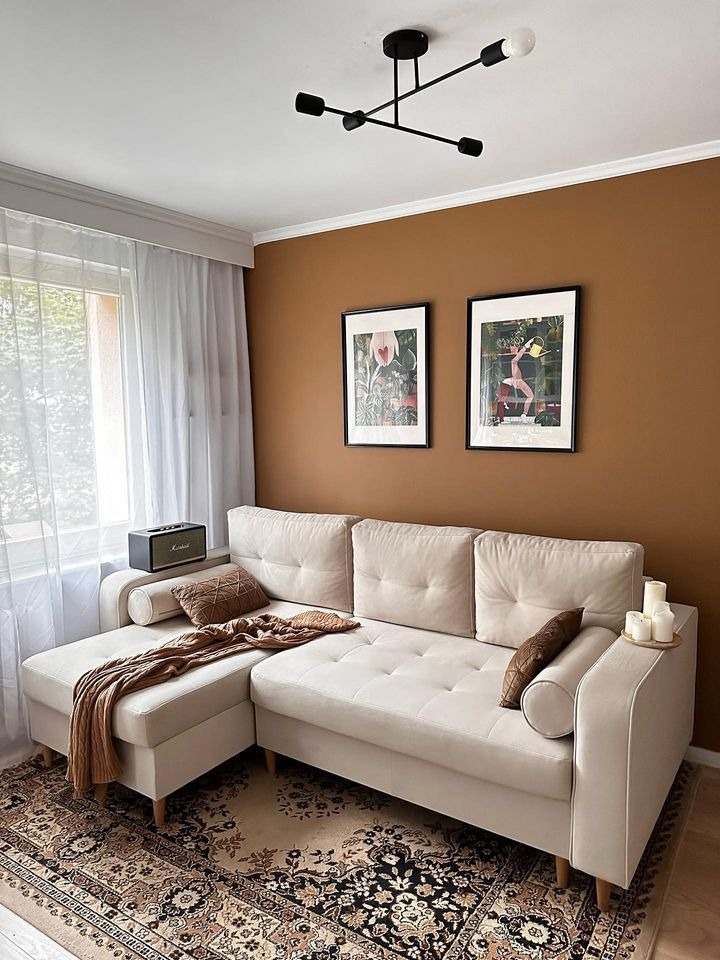 Beżowy narożnik z pikowaniem na tle miedzianej ściany, wzorzysty dywan, grafiki w czarnych ramkach i nowoczesna lampa sufitowa.