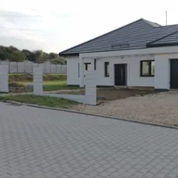 Wznoszenie ogrodzenia z prefabrykat&oacute;w betonowych przy nowoczesnym domu z garażem. Widoczne słupki i podmur&oacute;wka w trakcie montażu. Szara kostka brukowa na podjeździe.