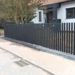 Nowoczesne, grafitowe ogrodzenie posesji z pionowych sztachet, z bramą wjazdową. U podstawy ogrodzenia szary kamień. Dom z numerem 76 w tle.