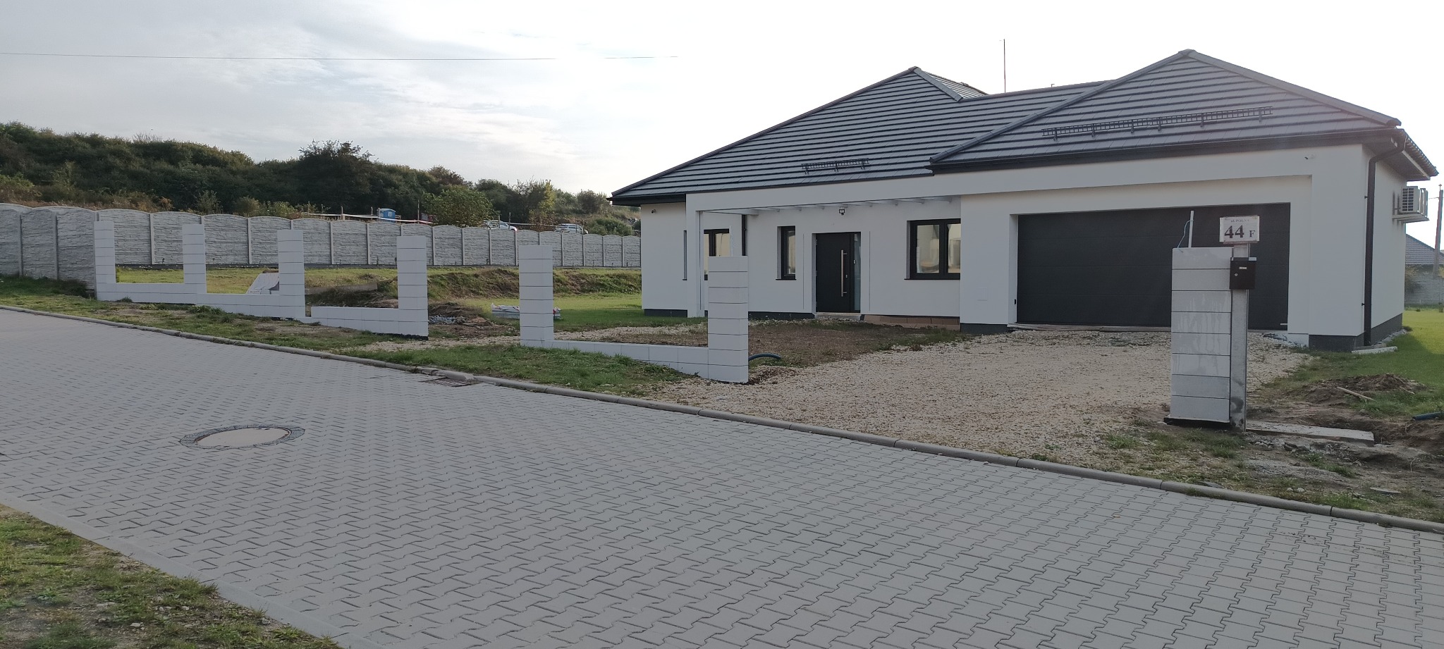 Wznoszenie ogrodzenia z prefabrykatów betonowych przy nowoczesnym domu z garażem. Widoczne słupki i podmurówka w trakcie montażu. Szara kostka brukowa na podjeździe.