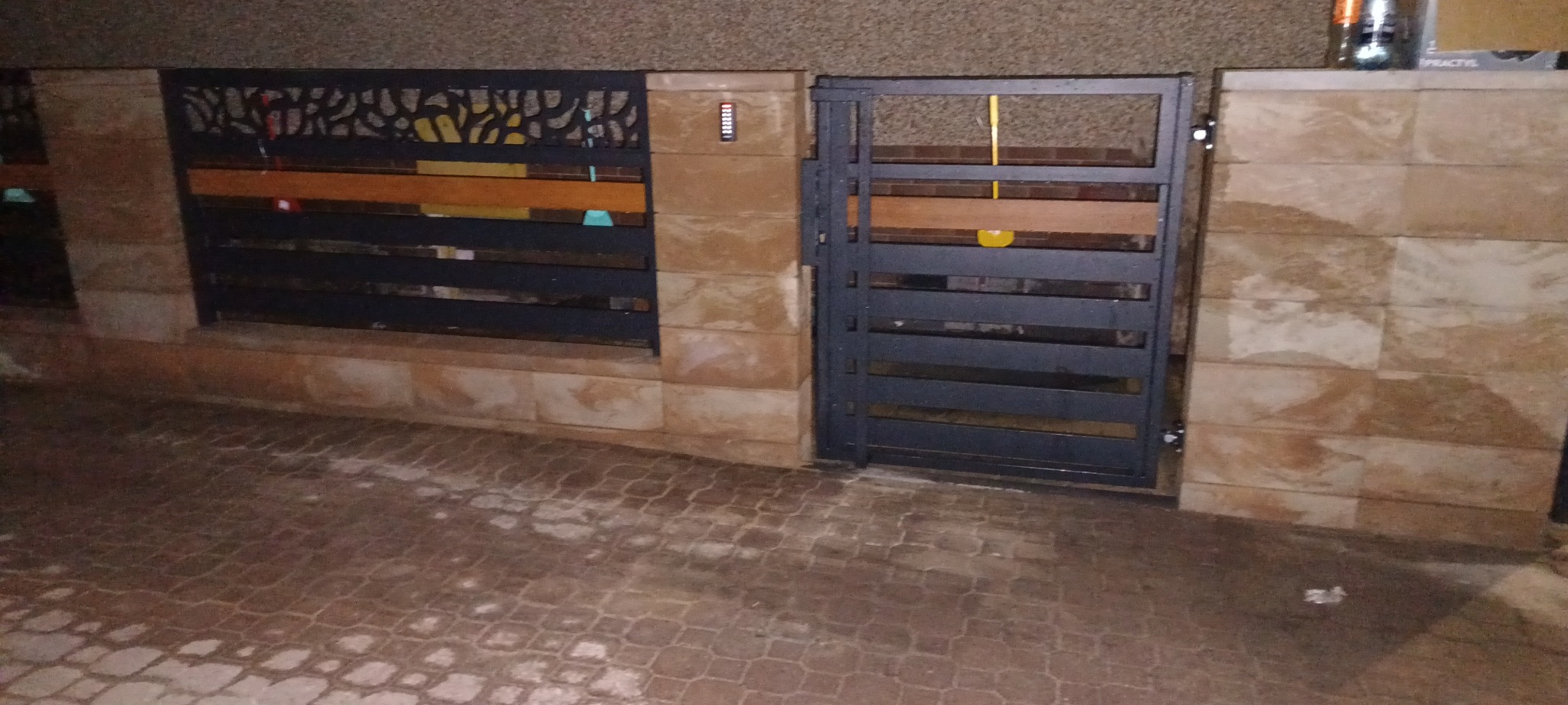 Nowoczesna brama wjazdowa z poziomymi panelami i elementami dekoracyjnymi, osadzona w murze z kamiennych bloków. Widoczne oświetlenie zewnętrzne.