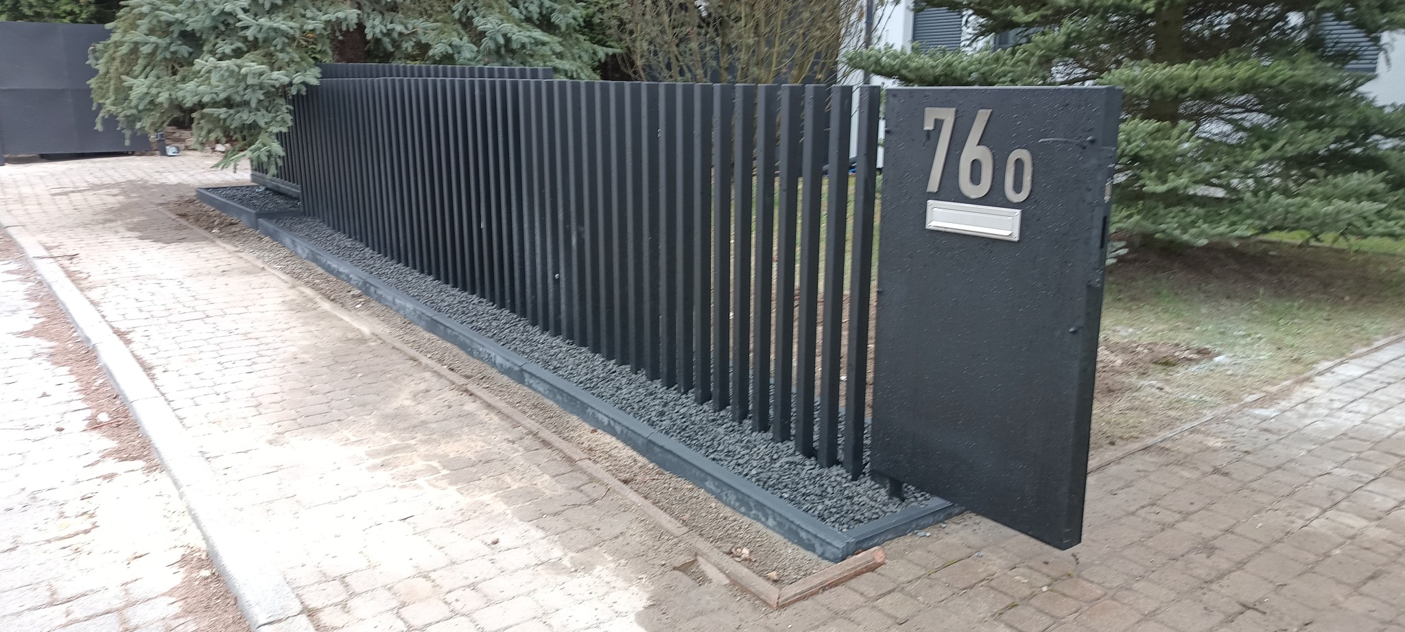 Nowoczesna, czarna brama wjazdowa z numerem domu '760' i skrzynką na listy, otoczona kamieniami dekoracyjnymi. Pionowe elementy bramy tworzą minimalistyczny design.