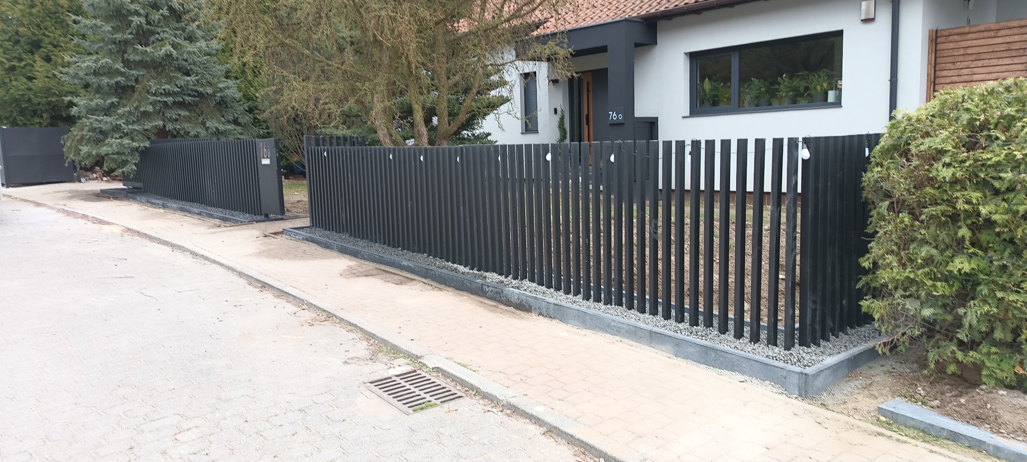 Nowoczesne, grafitowe ogrodzenie posesji z pionowych sztachet, z bramą wjazdową. U podstawy ogrodzenia szary kamień. Dom z numerem 76 w tle.