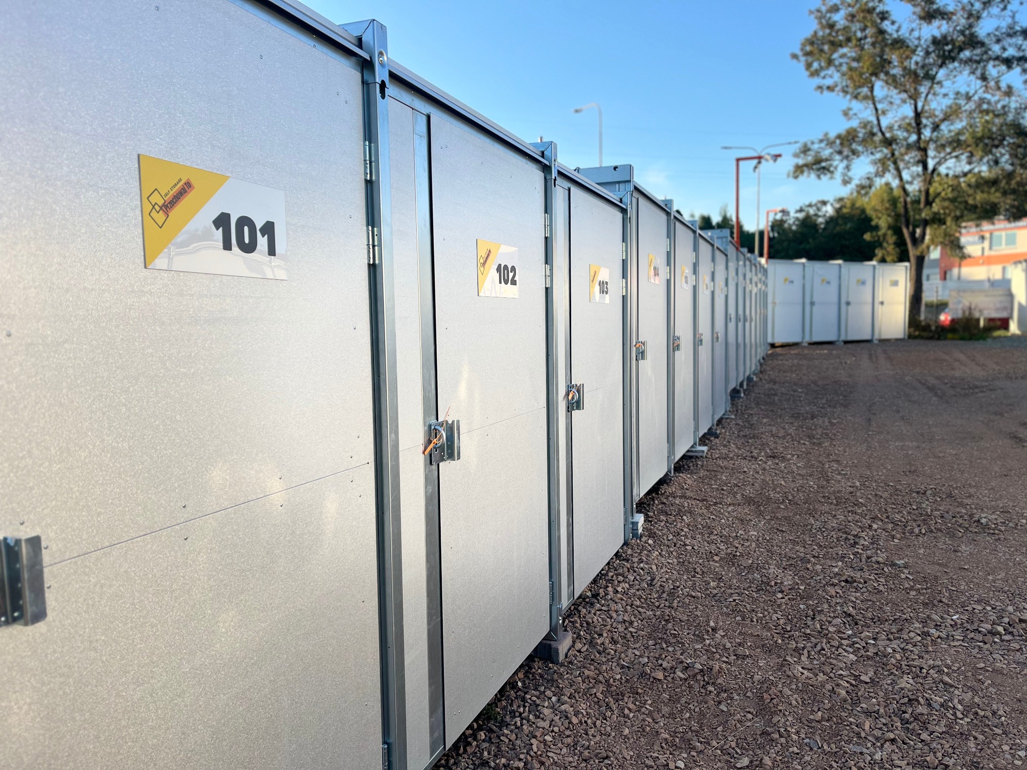 Rząd srebrnych, metalowych kontenerów magazynowych z numerami (101, 102, 103) na zewnątrz, na żwirowym podłożu, w tle drzewa i budynki.