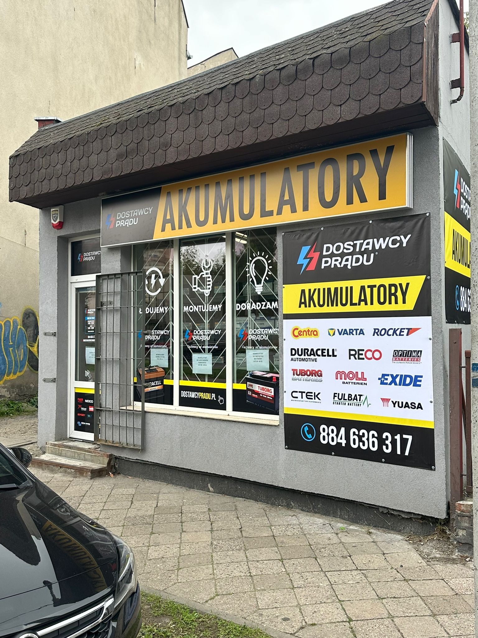 Fasada sklepu z akumulatorami 'Dostawcy Prądu' w Grudziądzu, z widocznym logo, numerem telefonu i ofertą montażu oraz doradztwa. Widoczne marki akumulatorów.
