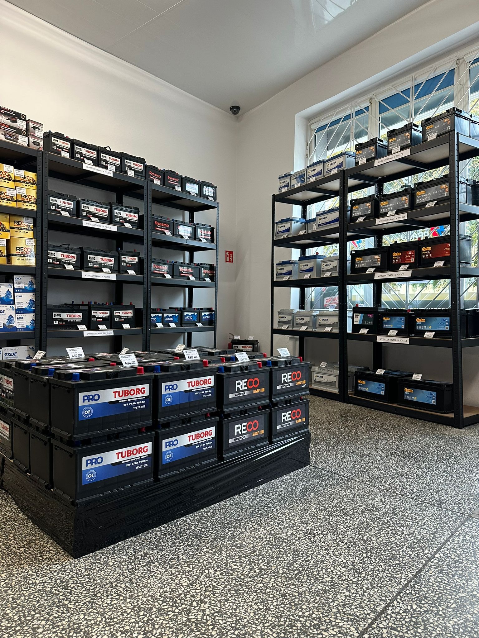 Sklep z akumulatorami: półki uginają się od Tuborg, Reco, Varta, Centra. Akumulatory ułożone na regałach i paletach, widoczny monitoring.