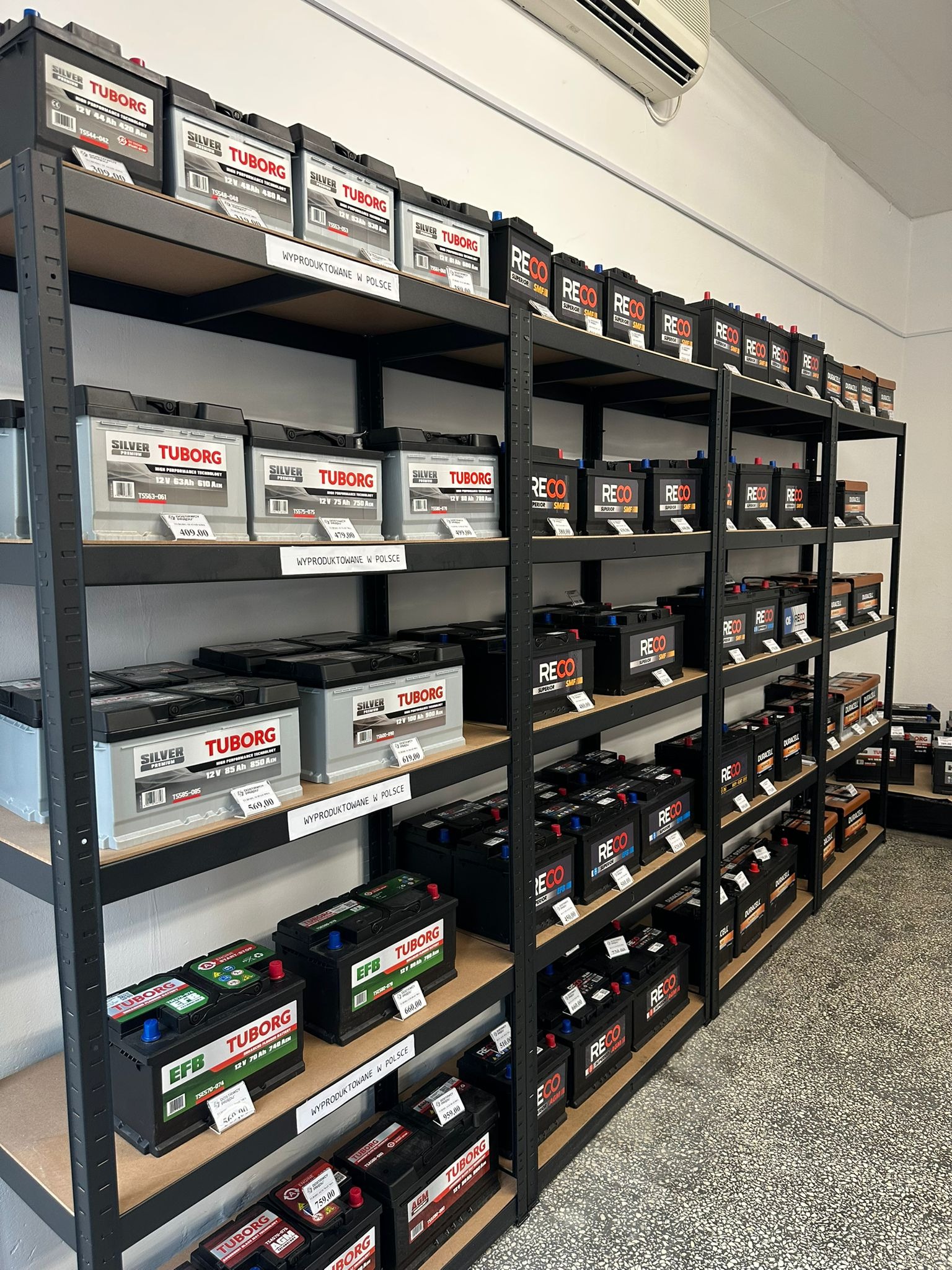 Regał sklepowy pełen akumulatorów samochodowych marek Tuborg, Reco, Varta, Centra, Dynamix, Arctic, Duracell, Exide, ułożonych na półkach. Widoczne ceny i napisy 'Wyprodukowano w Polsce'.