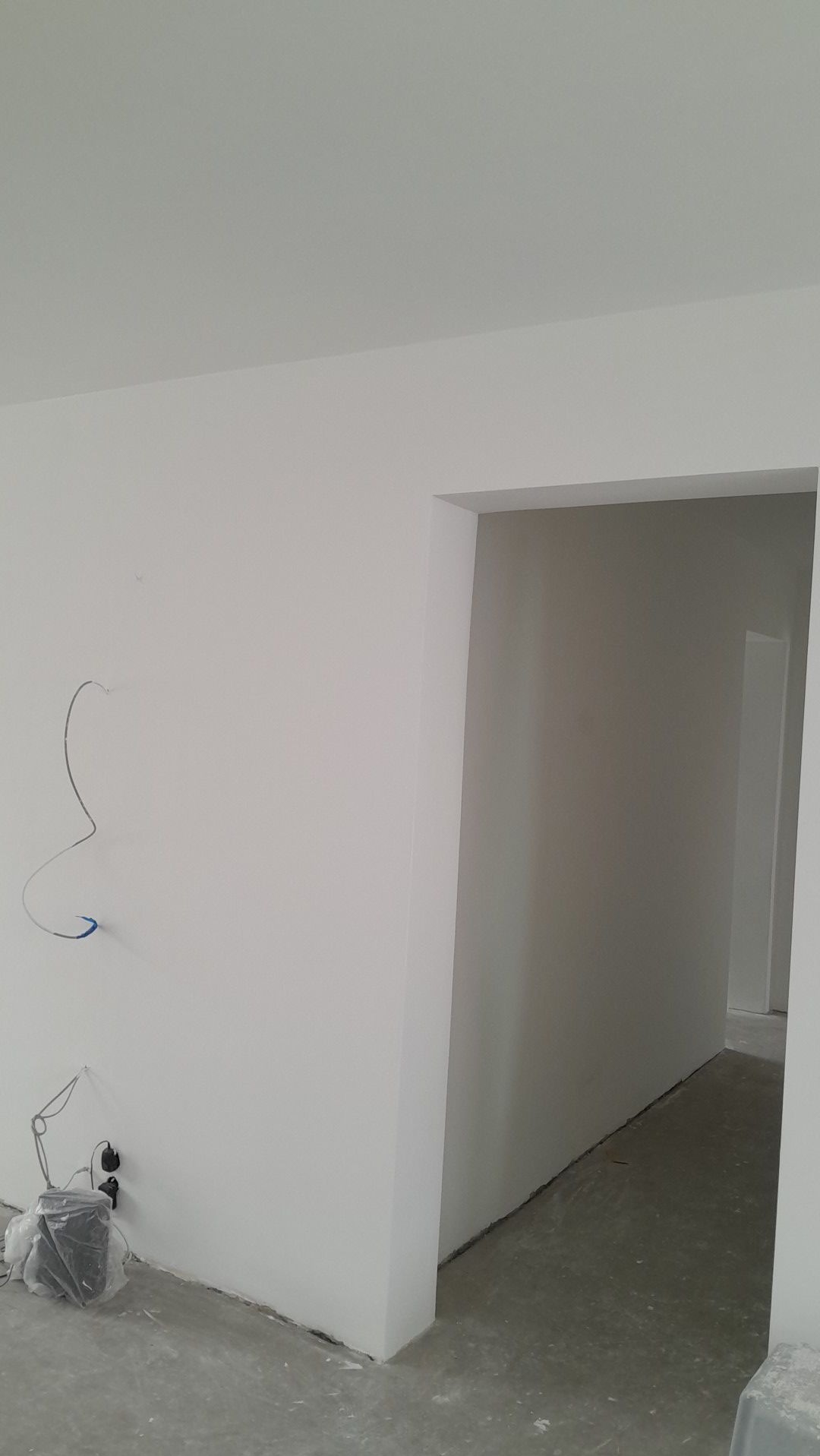 Wnętrze po remoncie: białe, gładkie ściany i sufit, widoczne kable elektryczne przy podłodze, surowy beton na podłodze, przejście do kolejnego pomieszczenia.