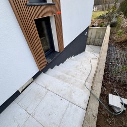 Daydrems Damian Hula - Nowoczesne wykończenie zewnętrzne domu: biała elewacja z drewnianymi lamelami, schody i taras wyłożone jasnoszarą płytką. Widoczny kabel i fragment ogrodu.