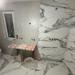 Daydrems Damian Hula - Łazienka w trakcie wykańczania w Poznaniu. Białe płytki imitujące marmur na ścianach i podłodze. Widoczne przygotowane instalacje wodne i stelaż pod umywalkę.