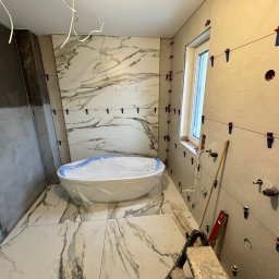 Daydrems Damian Hula - Łazienka w trakcie prac wykończeniowych: marmurowe płytki na podłodze i ścianach, wanna zabezpieczona folią, narzędzia glazurnicze, poziomica i niebieskie klipsy do płytek.