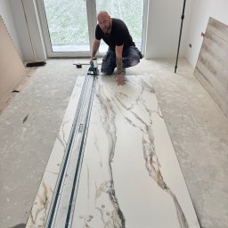 Daydrems Damian Hula - Glazurnik w trakcie pracy: docinanie płyty imitującej marmur przy użyciu specjalistycznej przecinarki. W tle widoczne okno i surowy stan wykończenia wnętrza.