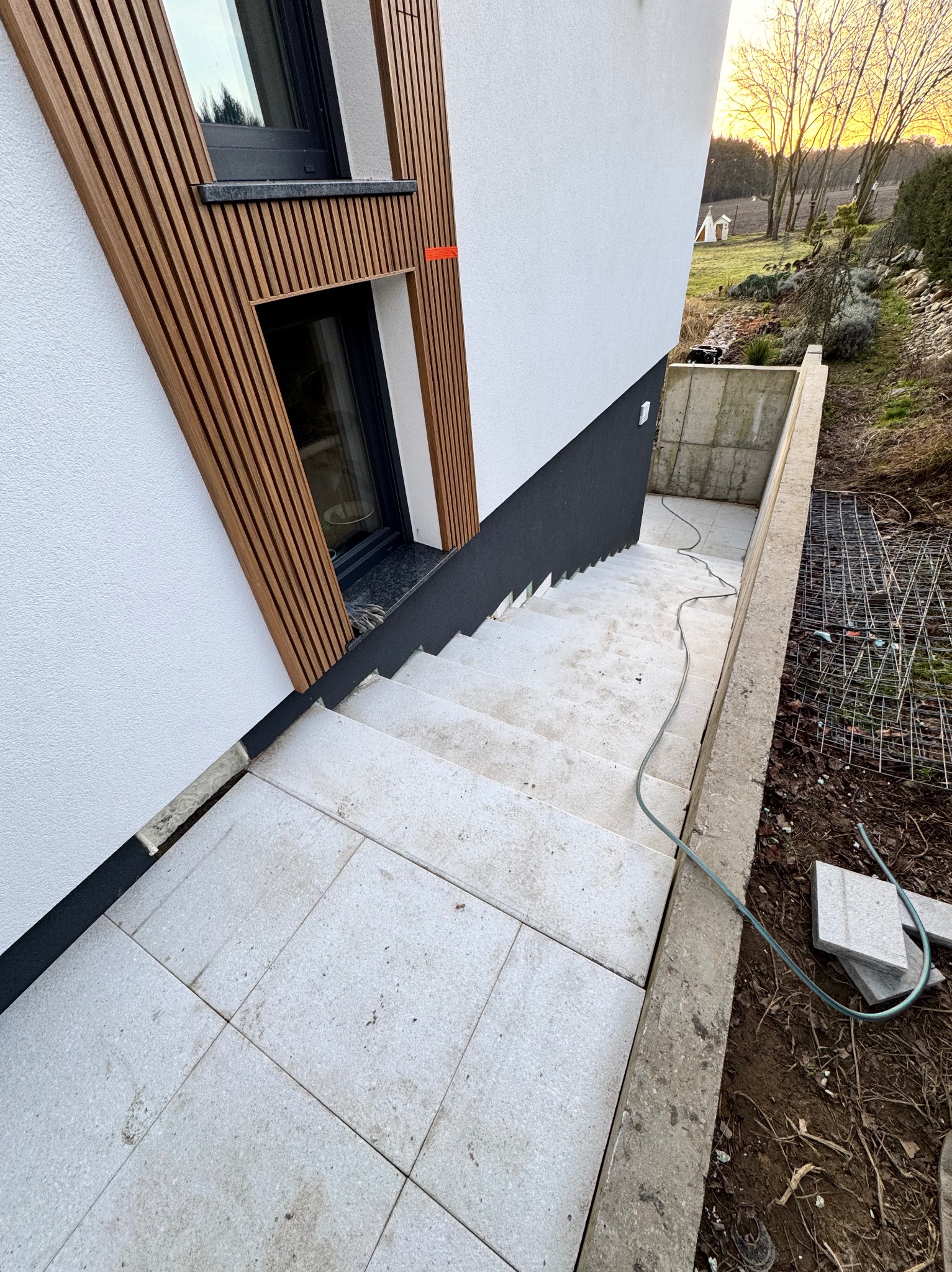 Nowoczesne wykończenie zewnętrzne domu: biała elewacja z drewnianymi lamelami, schody i taras wyłożone jasnoszarą płytką. Widoczny kabel i fragment ogrodu.