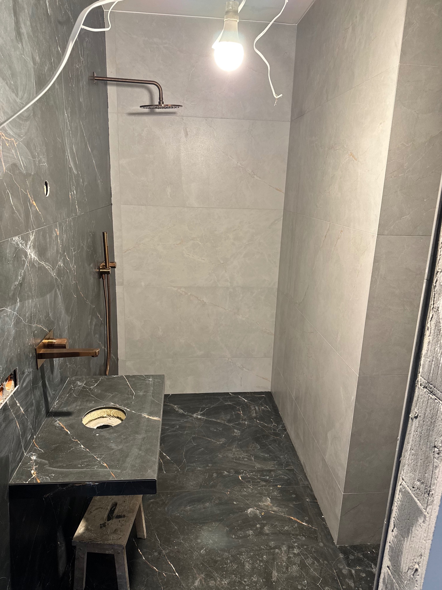Nowoczesna łazienka w trakcie wykańczania: marmurowe płytki, złota armatura, zamontowana umywalka. Widoczne przewody i taboret.