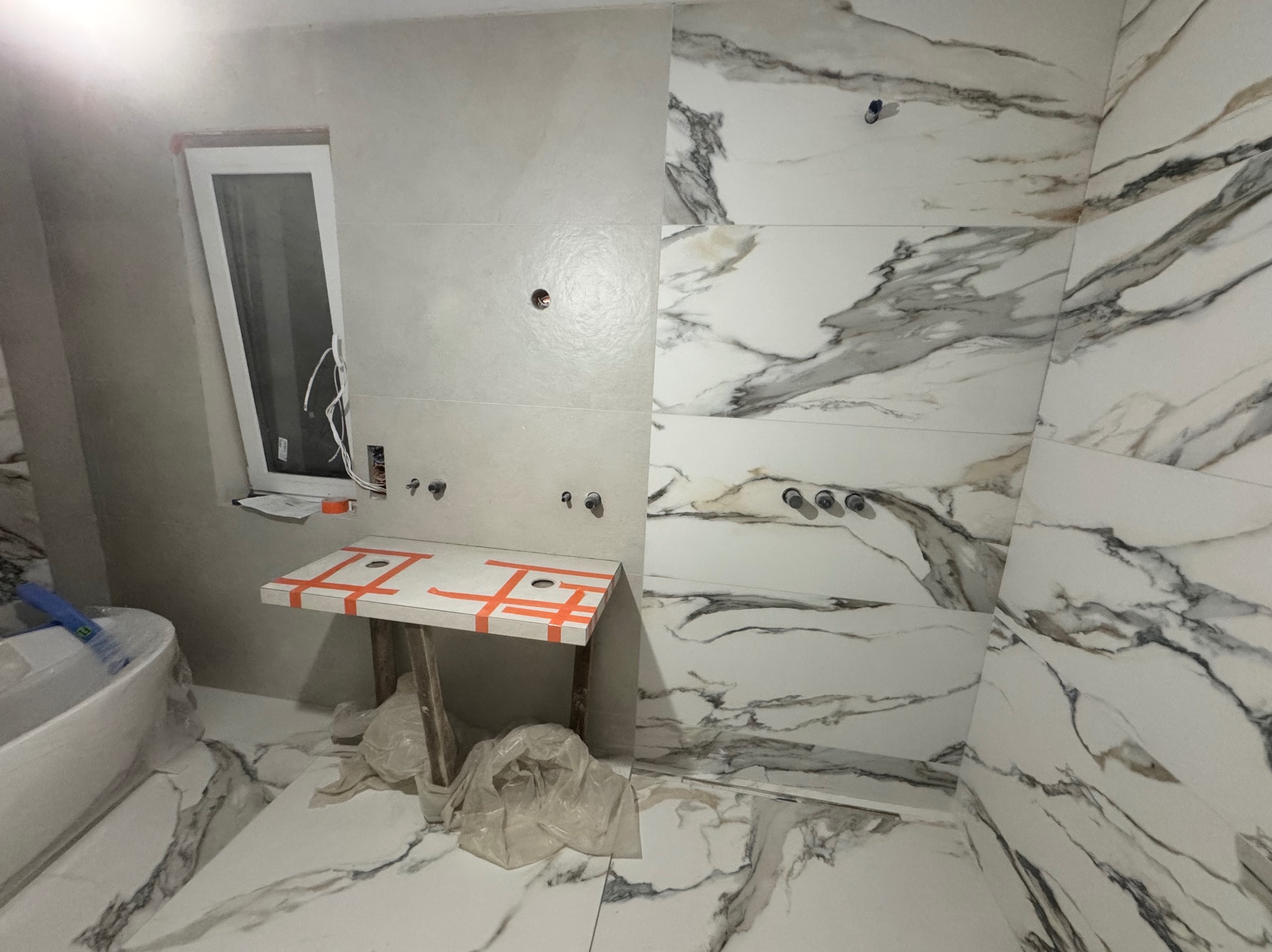 Łazienka w trakcie wykańczania w Poznaniu. Białe płytki imitujące marmur na ścianach i podłodze. Widoczne przygotowane instalacje wodne i stelaż pod umywalkę.