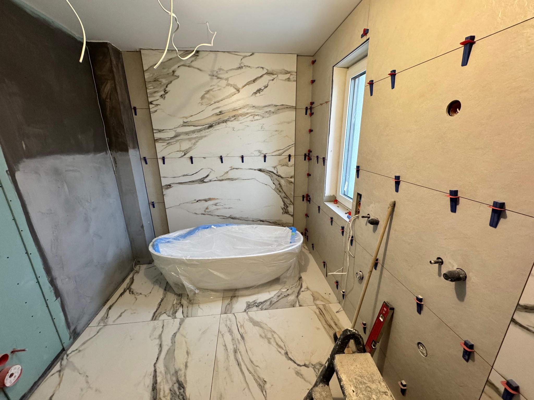 Łazienka w trakcie prac wykończeniowych: marmurowe płytki na podłodze i ścianach, wanna zabezpieczona folią, narzędzia glazurnicze, poziomica i niebieskie klipsy do płytek.