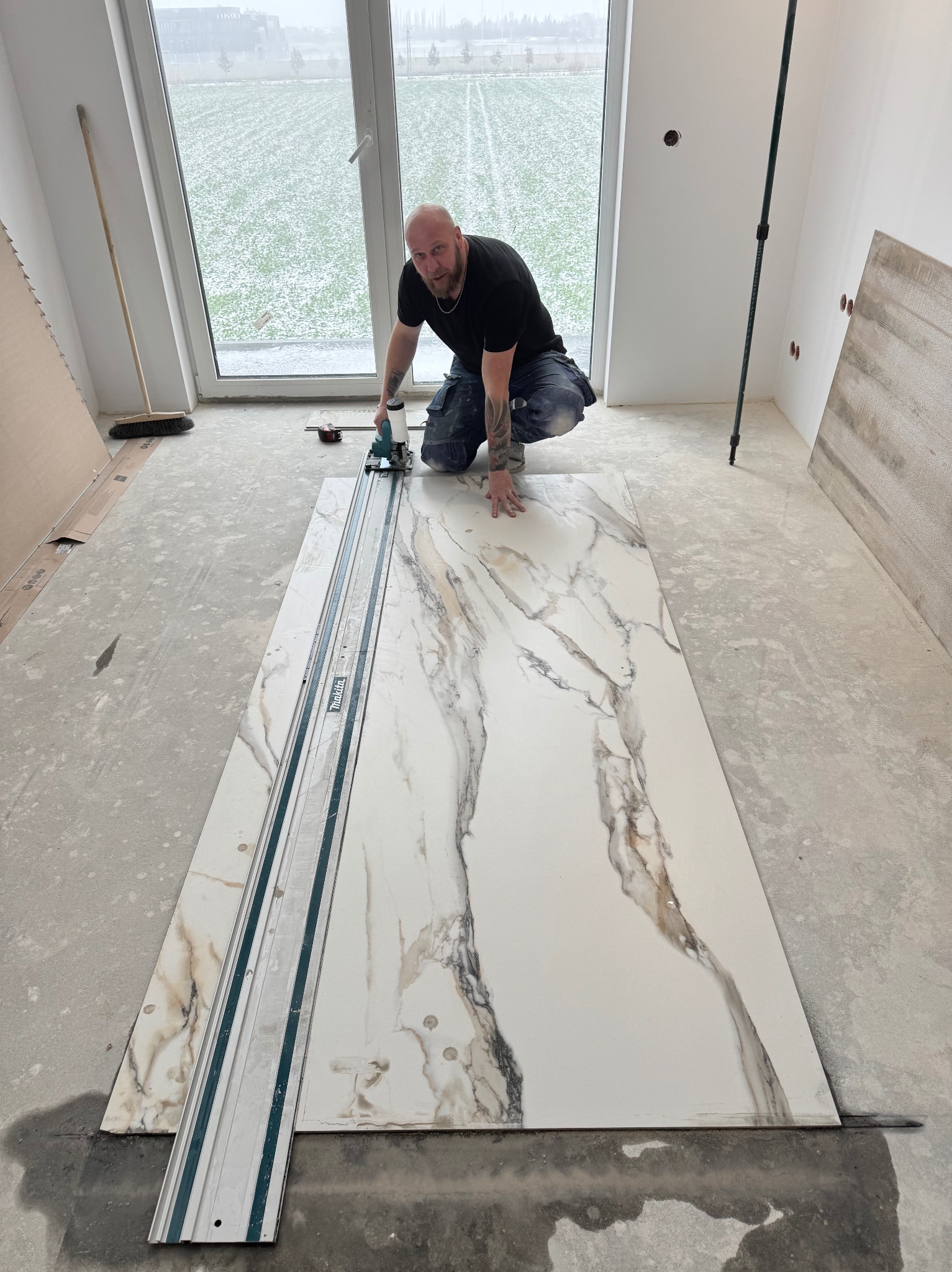 Glazurnik w trakcie pracy: docinanie płyty imitującej marmur przy użyciu specjalistycznej przecinarki. W tle widoczne okno i surowy stan wykończenia wnętrza.