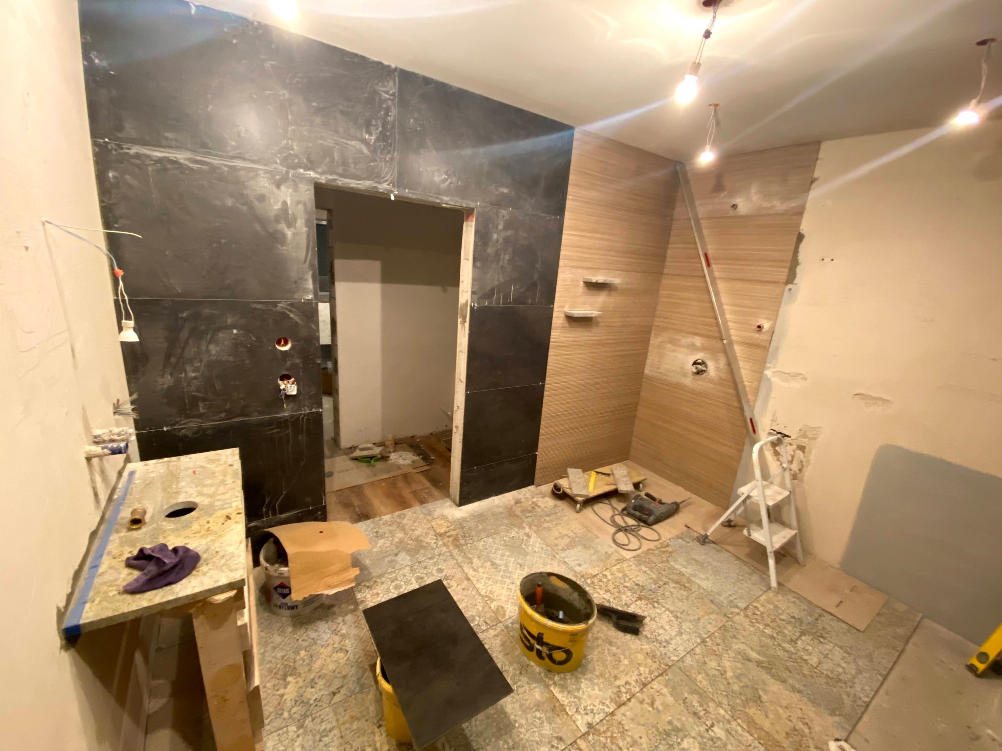 Wnętrze w trakcie prac wykończeniowych: ułożone płytki ścienne imitujące marmur i drewno, widoczne narzędzia glazurnicze i drabina. Fragment blatu z otworem.