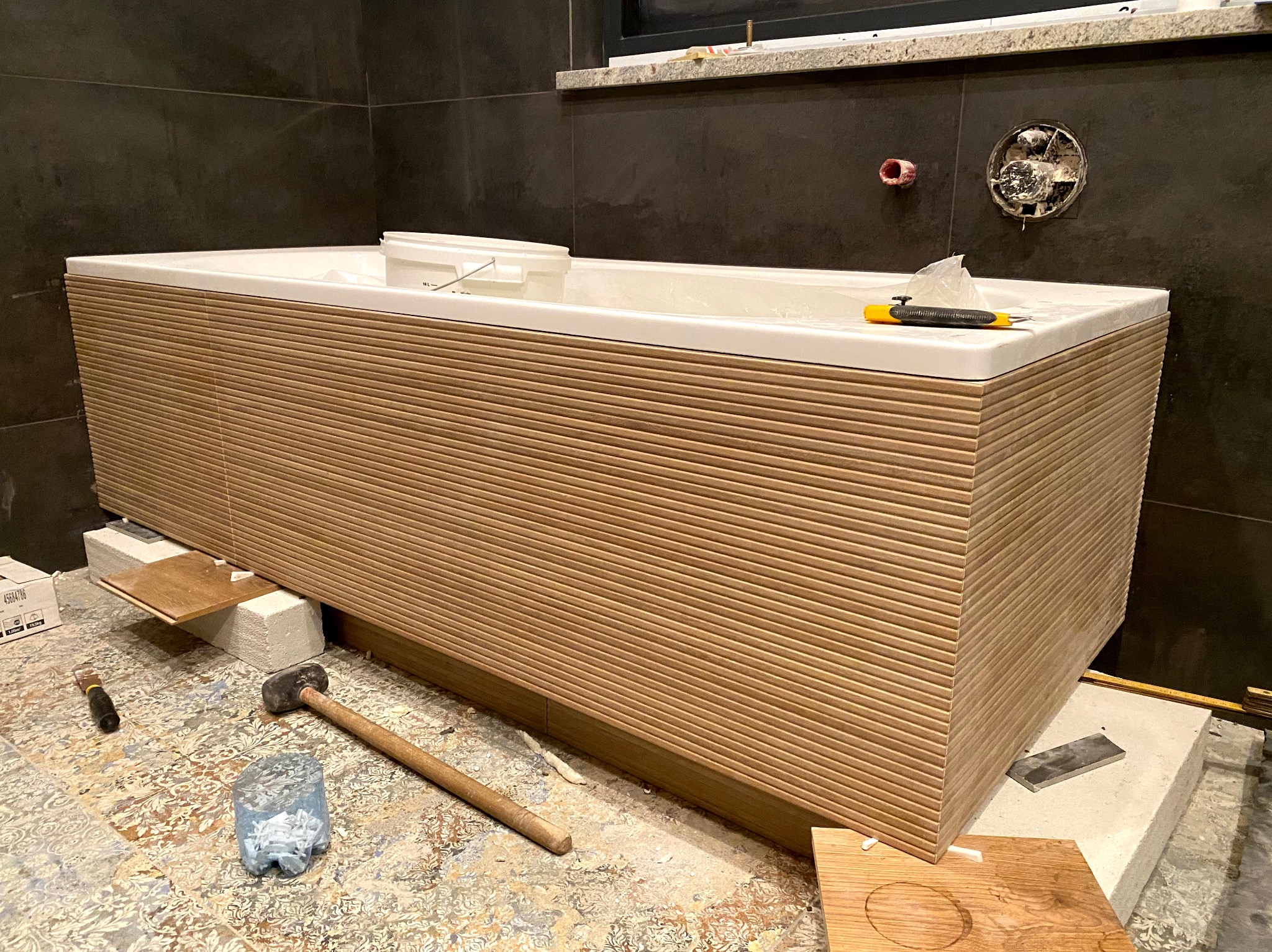 Biała wanna z dekoracyjną obudową w trakcie montażu w łazience z ciemnymi płytkami. Widoczne narzędzia i materiały budowlane.