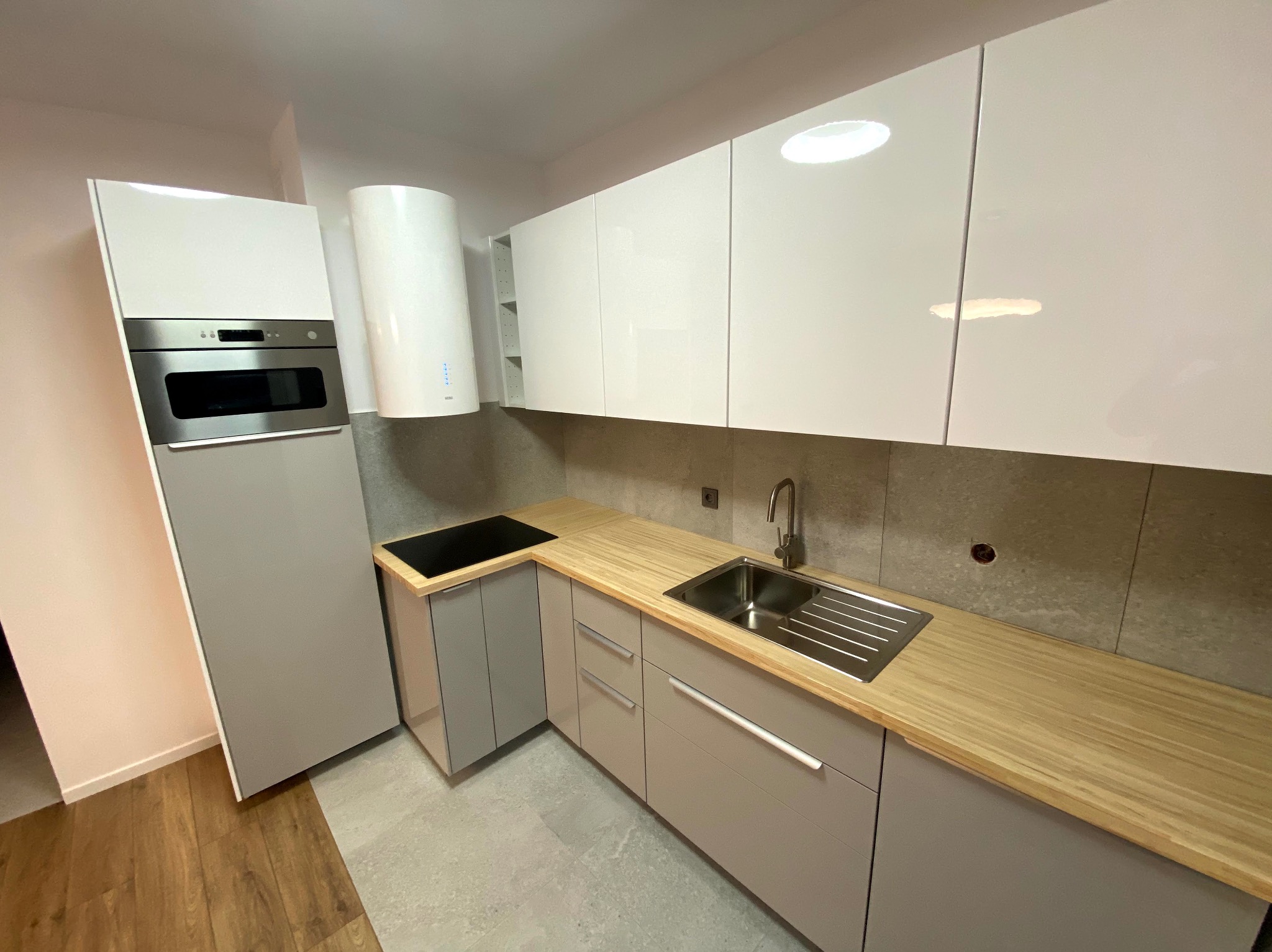 Nowoczesna kuchnia w odcieniach szarości i bieli z drewnianym blatem, widok na zlew, płytę indukcyjną i zabudowane sprzęty AGD. Minimalistyczny design.