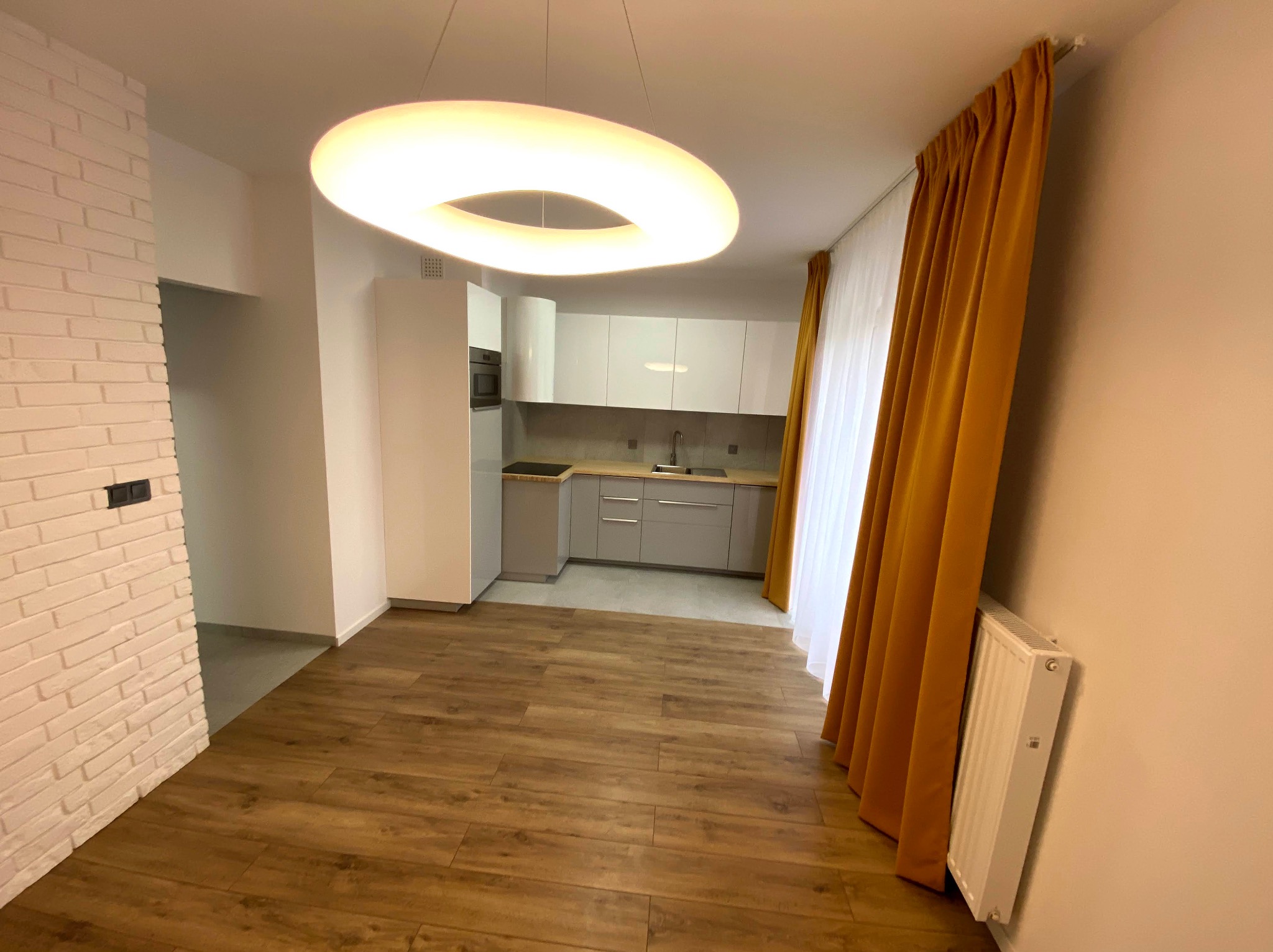 Jasne wnętrze po remoncie: drewnopodobna podłoga, biała cegła na ścianie, nowoczesna kuchnia z szarymi szafkami i okrągła lampa sufitowa.