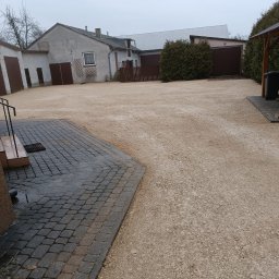 F.U. Kop-bud - Utwardzony teren żwirowy przed budynkiem z kostką brukową, widoczne ślady opon, szara pogoda.