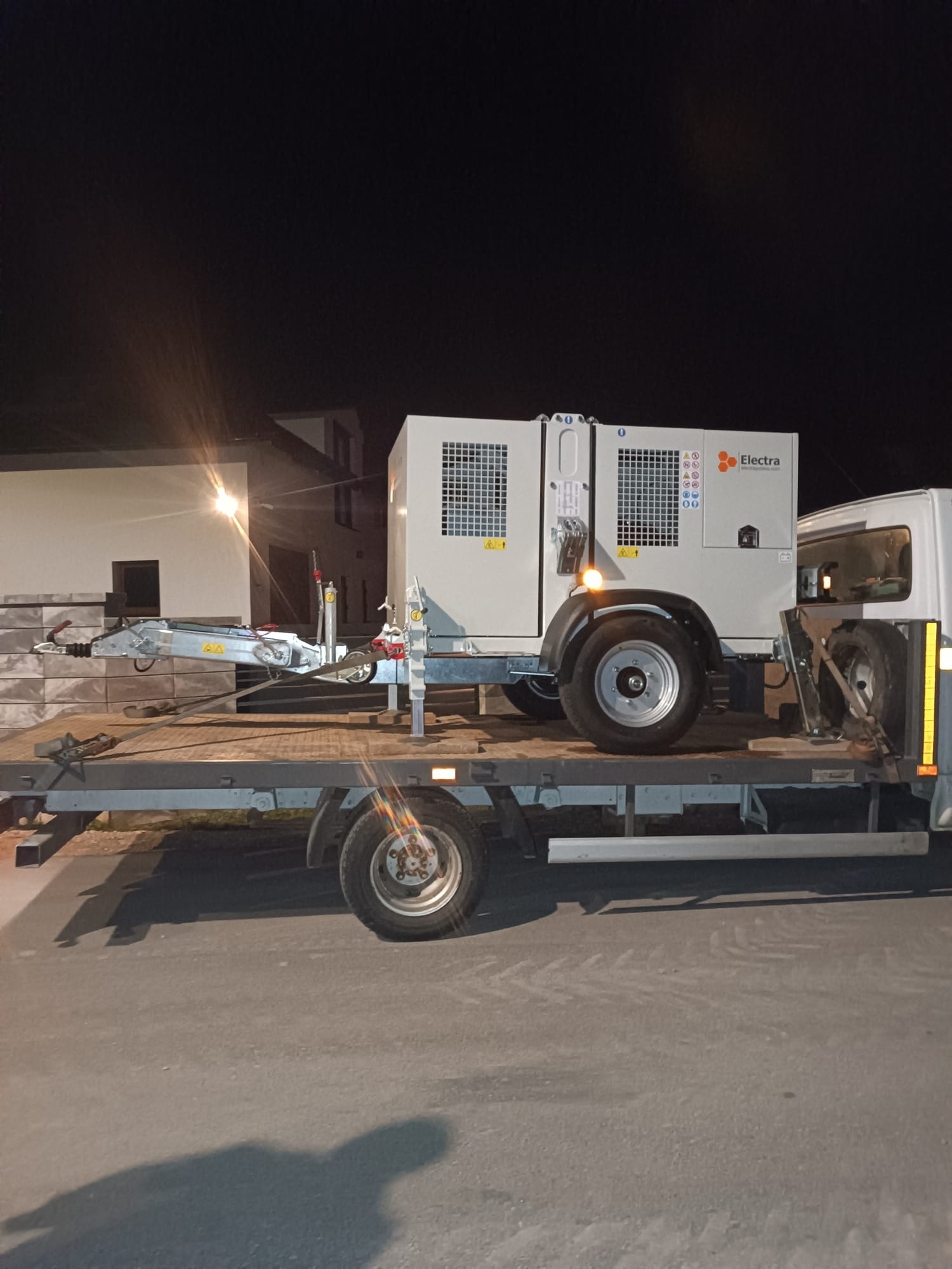 Generator Electra na przyczepie transportowej w nocy. Widoczny budynek w tle. Sprzęt gotowy do użycia przy niwelacji lub wykopach.