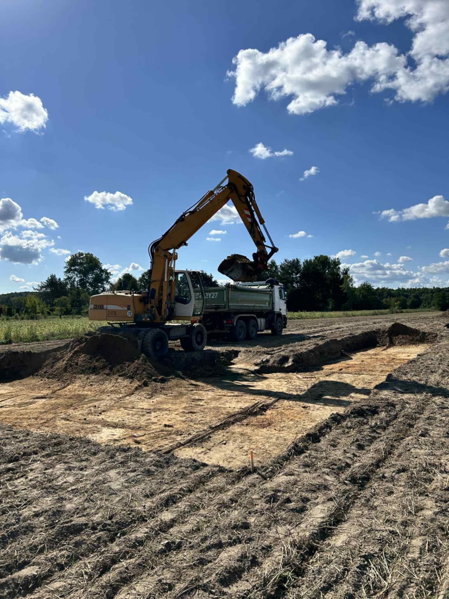 Żółta koparka ładuje ziemię na ciężarówkę podczas prac ziemnych w Głuchowie. Wykopane rowy w ziemi. Błękitne niebo z chmurami w tle.