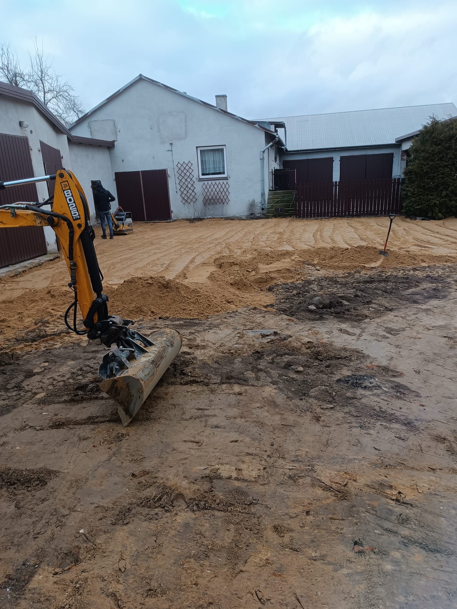Żółta koparka JCB na placu budowy, w tle budynek i pracownik używający zagęszczarki. Widoczne ślady opon i częściowo wyrównany teren.