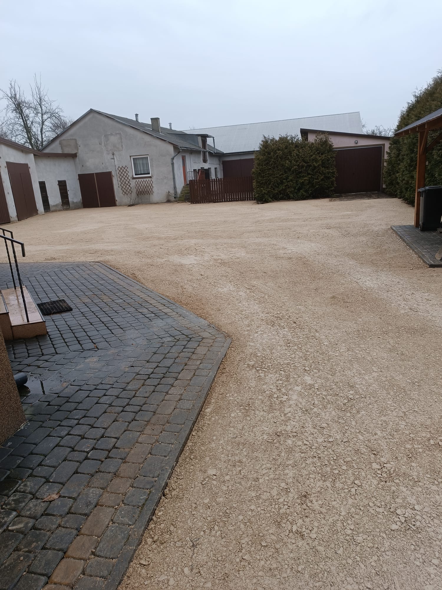 Utwardzony teren żwirowy przed budynkiem z kostką brukową, widoczne ślady opon, szara pogoda.