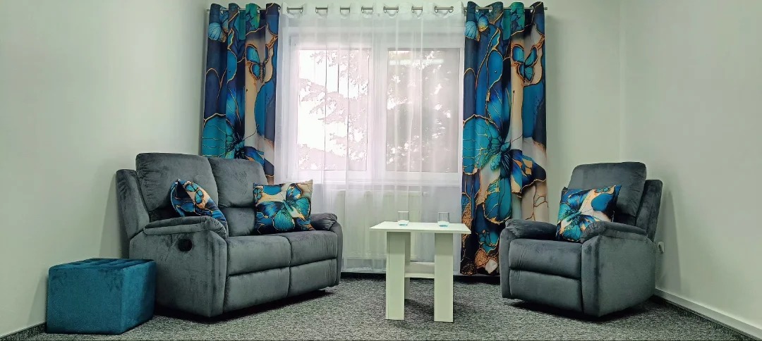 Przytulne wnętrze gabinetu: szara sofa i fotel z poduszkami w motyle, biały stolik z dwoma szklankami, okno z zasłonami w kwiaty, turkusowy puf.