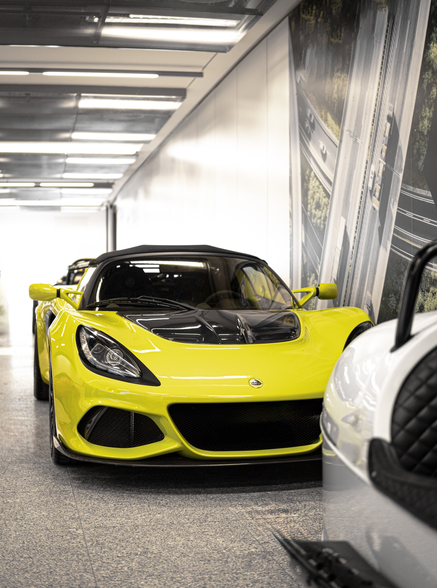 Żółty Lotus Elise parkuje w jasnym garażu z fototapetą autostrady. Widoczne detale: czarny grill, reflektory i logo Lotus. Część białego auta po prawej.