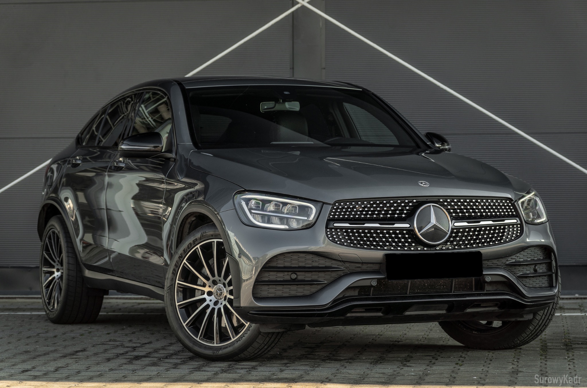 Szary Mercedes GLC Coupe na tle szarej ściany z białymi liniami. Widok z przodu, skupiony na detalu grilla i reflektorów. Efektowne felgi i opony.