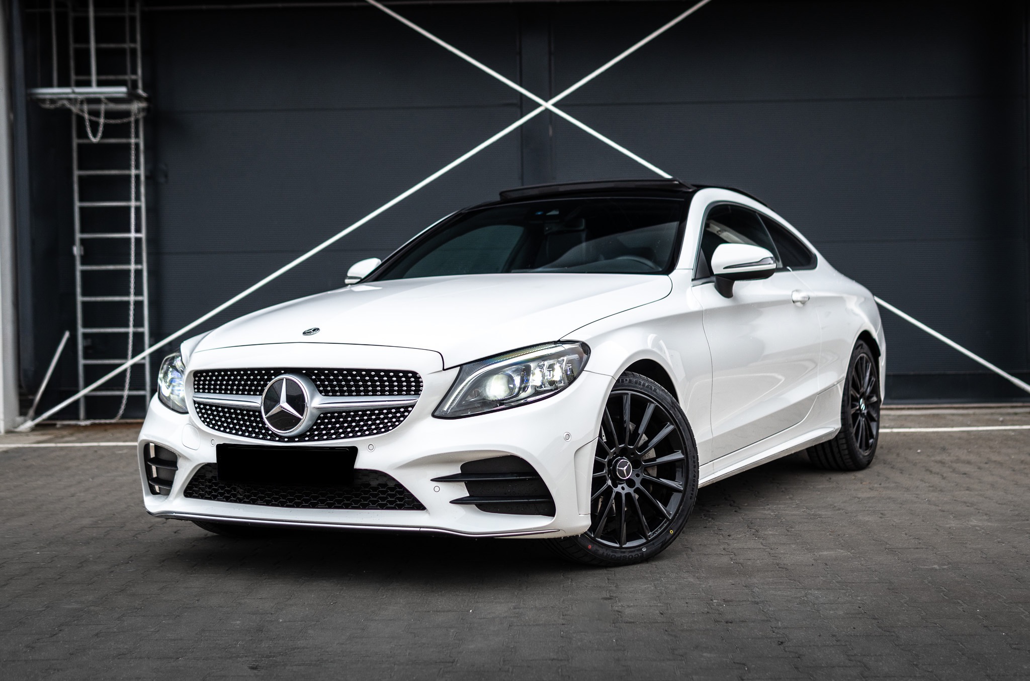 Białe coupe Mercedes-Benz C-klasy z czarnymi felgami na tle szarej ściany z białymi liniami. Auto zaparkowane na szarym bruku, widoczne detale.