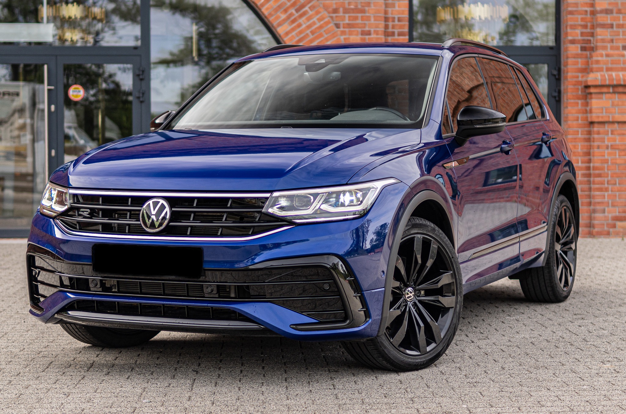 Błyszczący, ciemnoniebieski Volkswagen Tiguan R-Line z czarnymi felgami na tle ceglanej ściany i szklanych drzwi wejściowych. Precyzyjne detale i głębia koloru.