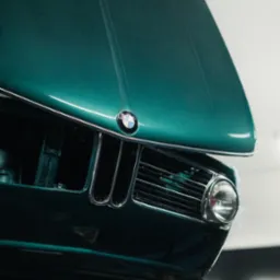 Zielony, klasyczny BMW z otwartą maską, widok z dołu, eksponujący atrapę chłodnicy i reflektor. Widoczne detale karoserii i logo marki.