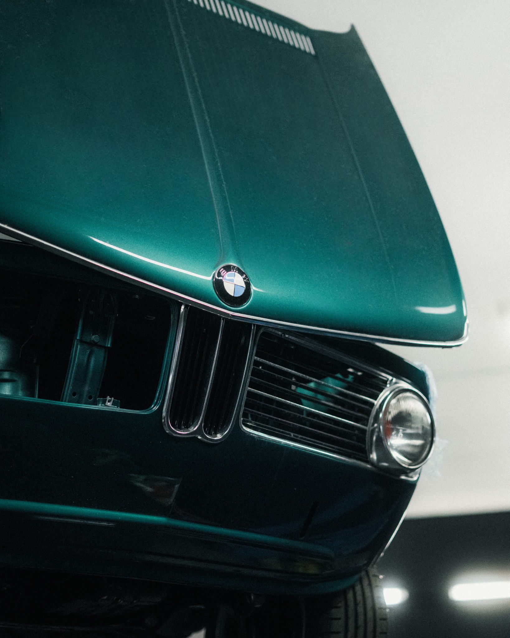 Zielony, klasyczny BMW z otwartą maską, widok z dołu, eksponujący atrapę chłodnicy i reflektor. Widoczne detale karoserii i logo marki.