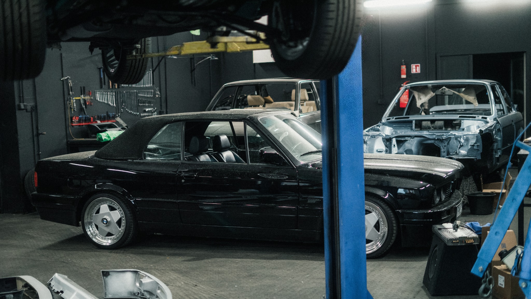 Warsztat samochodowy: czarny kabriolet BMW E30 na pierwszym planie, w tle inne pojazdy w trakcie naprawy, podnośnik i narzędzia.