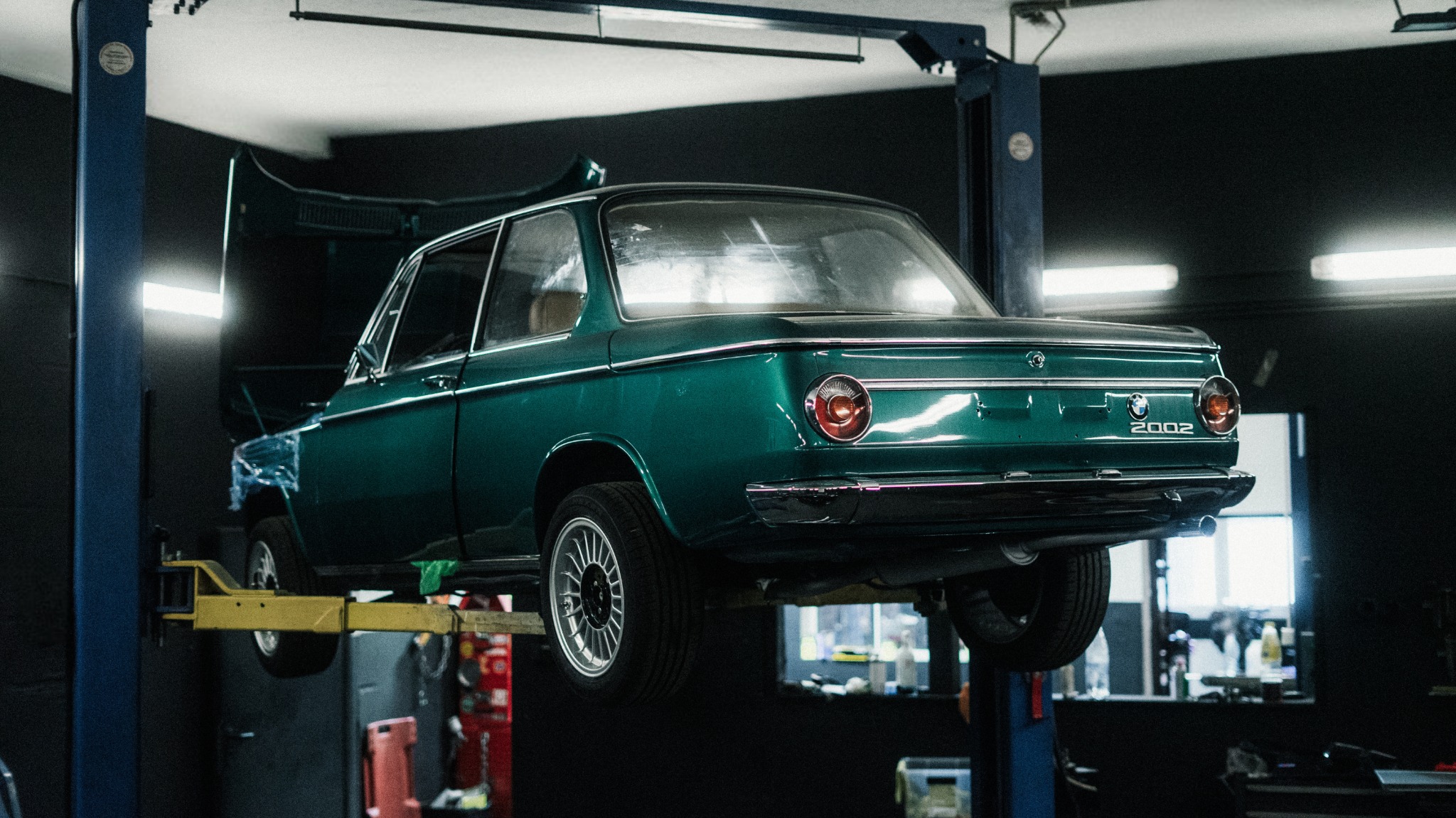 Zielone BMW 2002 na podnośniku w warsztacie. Otwarta maska, widoczne tylne światło i tabliczka z modelem. Ciemne tło, oświetlenie warsztatowe.