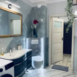 Elegancka łazienka z marmurowymi płytkami, umywalką z szafką w kolorze butelkowej zieleni i złotym lustrem. Oświetlenie LED podkreśla nowoczesny design.