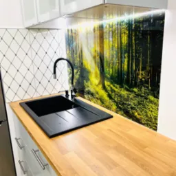 Nowoczesna kuchnia: czarny zlew z baterią, drewniany blat, dekoracyjny panel ścienny z motywem leśnym i białe szafki z chromowanymi uchwytami.